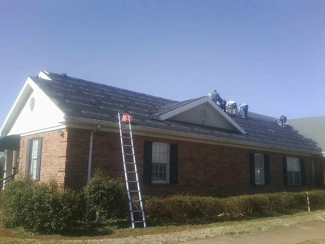 Crew tearing shingles off a brick ranch roof with a ladder set — Emergency Roof Repair work by our crew in Glen Campbell