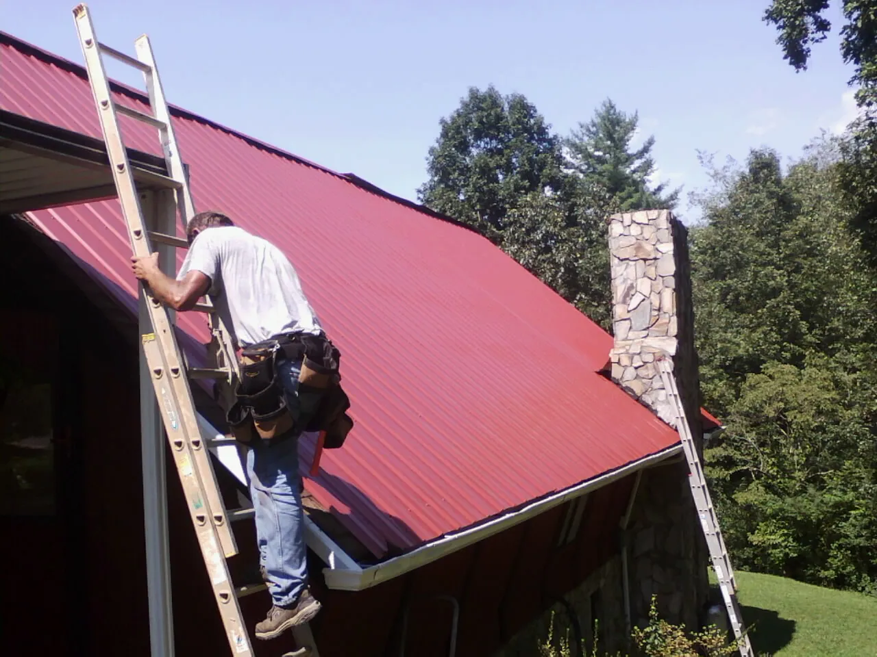 Roofer on a ladder beside a red metal roof and stone chimney — Soffit & Fascia Repair by Roofing Brothers in Glen Campbell